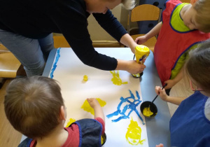 Na stoliku leży karton częściowo pomalowany farbami. Koło stolika stoi troje dzieci w fartuszkach. Nad stolikiem pochyla się nauczycielka. Wyciska z butelki farbę do małej miseczki.