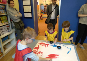 Na stoliku leży karton pomalowany częściowo farbami. Przy stoliku stoi troje dzieci. Trzymają pędzle i malują na kartonie. W tle widać stojących mężczyznę i kobietę. Za nimi widać regały z pólkami i wejście do sali.
