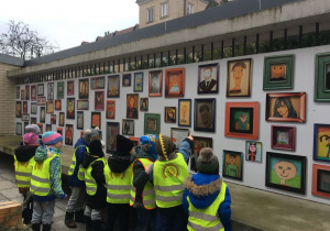 Dzieci w kamizelkach odblaskowych stoją tyłem przed tablicą. Tablica znajduje się na ogrodzeniu budynku. Na niej przyczepione są portrety osób w ramkach. Dzieci rękami wskazują portrety.