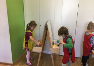 Dwoje dzieci stoi naprzeciwko siebie. Między nimi ustawiona jest sztaluga. Dzieci malują na sztaludze. Z tyłu widać białe drzwi szaf. Po prawej stronie widać stojącą dziewczynkę.