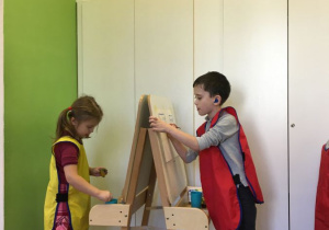 Dwoje dzieci stoi naprzeciwko siebie. Między nimi ustawiona jest sztaluga. Dzieci malują na sztaludze. Z tyłu widać białe drzwi szaf i fragment zielonej ściany.