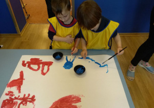 Na stoiku leży duży karton. Na nim namalowane są niebieską i czerwona farbą kształty. Za stolikiem stoi dwóch chłopców w żółtych fartuszkach. Chłopcy trzymają pędzle i malują na kartonie niebieską farbą.