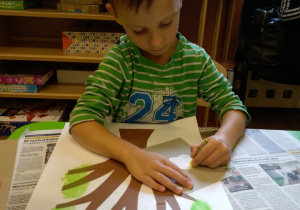 Przy stole siedzi chłopiec. Na stole przed nim leży kartka z wyklejonym z papieru drzewem. Chłopiec trzyma w ręku kredkę. Rysuje na drzewie liście. W tle widać okno i regał z półkami.