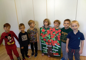 Grupa dzieci stoi w półkolu na tle białych drzwi. Dwoje dzieci trzyma przed sobą duży karton papieru. Na kartonie wyklejone jest drzewo z czerwonymi jabłkami.