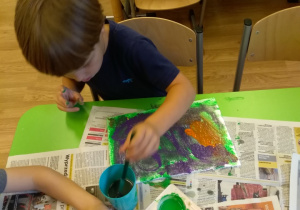 Chłopiec siedzi przy stole. Na stole rozłożone są gazety. Na nich leży kartka pomalowana farami. Chłopiec wkłada pędzel do kubka. Obok stoi pojemnik z farbami.