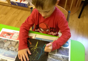 Przy stole siedzi chłopiec. Przed nim na gazetach leży kartka pokryta czarną farbą. Chłopiec trzyma w ręku patyczek i rysuje nim po kartce.