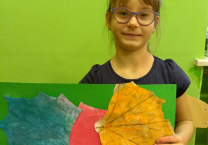 Na tle zielonej ściany stoi dziewczynka. Trzyma przed sobą zieloną kartkę. Na kartce naklejone są trzy liście wycięte z papieru. Liście pokolorowane są niebiesko, różowo i pomarańczowo.