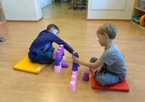 Na podłodze na poduszkach siedzą naprzeciwko siebie dwaj chłopcy. Każdy chłopiec buduje wieżę z kolorowych kubków: różowych, granatowych i fioletowych.
