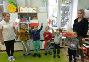 Dzieci i dwie osoby dorosłe stoją w szeregu. Dzieci trzymają w rękach biało-czerwone chorągiewki. Unoszą je do góry. W tle widać okno i półkę z zabawkami.