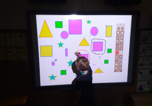 Przed tablicą multimedialną stoi chłopiec. Na tablicy wyświetlone są kolorowe figury geometryczne. Chłopiec trzyma w ręku flamaster i obrysowuje jedną z figur.