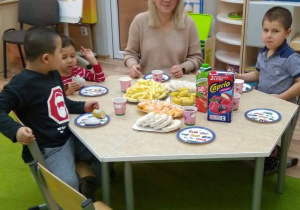 Przy stoliku siedzi dyrektor przedszkola i trzech chłopców. Na stole stoją kubki, talerze i miski z owocami i chrupkami. Z tyłu widać drzwi i meble.