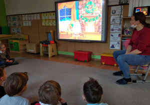Na tablicy multimedialnej wyświetlany jest film. Po lewej stronie na krześle siedzi nauczycielka. Patrzy na tablice. Z przodu widać głowy dzieci.