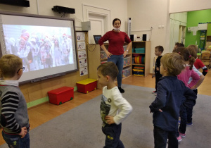 Nauczycielka stoi z rękami na biodrach. Po jej prawej stronie jest tablica multimedialna. Pod tablicą stoją dwa czerwone pojemniki. Na tablicy wyświetlany jest film. Dzieci stoją w półkolu. Są zwrócone twarzami do nauczycielki. Ręce trzymają na biodrach.