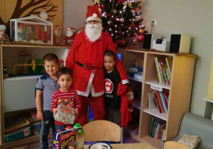 Postać w stroju Świętego Mikołaja stoi na tle półek z zabawkami. Przy postaci stoi trzech chłopców. Dwóch po prawej, jeden po lewej. Chłopiec po lewej stronie przytula się do Mikołaja.