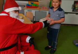 Osoba w stroju Świętego Mikołaja siedzi na krześle. Przed nią stoi chłopiec. Postać podaje prezent dziecku. W tle widać okno i meble.