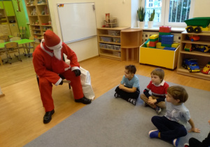 Osoba w stroju Świętego Mikołaja siedzi na krześle. Pochyla się do przodu. Jeną rękę opiera o kolano. Po prawej stronie na dywanie siedzi trzech chłopców. Patrzą na Mikołaja.