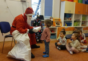 Osoba w stroju Świętego Mikołaja siedzi na krześle. W rękach trzyma prezent. Przed nią stoi dziecko. Przy krześle stoi worek. Po lewej stronie na dywanie siedzą dzieci. W tle widać choinkę, okno i meble.