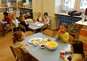 W sali przy stolikach siedzą dzieci. Na stolikach stoją miski i talerze z chrupkami, bananami i waflami ryżowymi. Część dzieci trzyma w rękach papierowe kubki. W tle widać meble i okna.