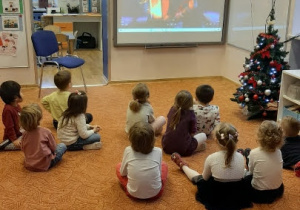 Przed tablicą multimedialną w luźnej gromadce siedzą dzieci. Patrzą na film wyświetlany na tablicy.