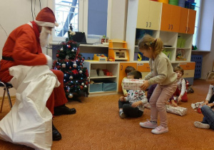 Osoba w stroju Świętego Mikołaja siedzi na krześle. Jedną ręką przytrzymuje worek. Przed nią stoi dziewczynka z prezentem w rękach. Za dziewczynką na dywanie siedzą dzieci. W tle widać choinkę, okno i meble.
