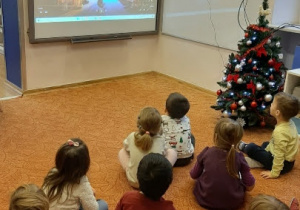 Przed tablicą multimedialną w luźnej gromadce siedzą dzieci. Patrzą na film wyświetlany na tablicy. Pop lewej stronie stoi choinka.