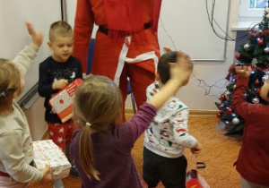 Postać w stroju Świętego Mikołaja stoi z podniesioną ręką. Wokół niej stoją dzieci. Niektóre mają podniesione ręce.