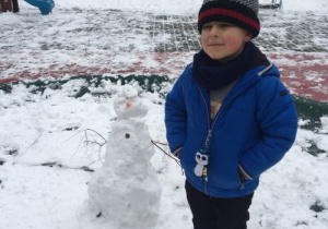 Chłopiec w zimowym ubraniu stoi na podwórku przedszkolnym. Na ziemi leży warstwa śniegu. Po prawej stronie chłopca stoi mały bałwan.