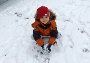 Chłopiec w zimowym ubraniu przykucnął i patrzy przed siebie. W rękach trzyma śnieg. Dookoła niego leży warstwa śniegu.