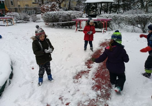 Grupa dzieci w zimowych ubraniach stoi na śniegu. W rękach trzymają kulki śniegu. Za nimi widać drzewa i domek ogrodowy pokryte śniegiem.