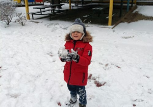 Chłopiec w zimowym ubraniu stoi na podwórku przedszkolnym. W rękach trzyma kulkę śniegu. Na ziemi leży warstwa śniegu. Za chłopcem widać wiatę ogrodową i drewniany pociąg.