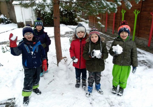 Grupa dzieci w zimowych ubraniach stoi na podwórku. W rękach trzymają kulki śniegu. Za nimi widać fragment drzewa i domku ogrodwoego.