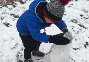 Chłopiec w zimowym ubraniu pochyla się i rękami dotyka bałwana. dookoła leży warstwa śniegu.