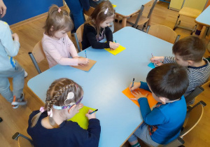Przy stoiku siedzi pięcioro dzieci. Przed nimi leżą kolorowe kartki. Dzieci trzymają w rękach ołówki. Odrysowują na karkach swoje dłonie.