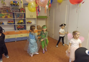 Dzieci w strojach karnawałowych stoją na dywanie. Dwie dziewczynki trzymają się za ręce. U góry widać fragmenty kolorowych balonów. Z tyłu stoi regał z samochodami i innymi zabawkami na półkach.