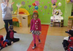 Na podłodze rozłożona jest długa czerwona tkanina. Po niej idzie dziewczynka w stroju karnawałowym. Po prawej stronie stoi nauczycielka. Klaszcze w dłonie. Po lewej i po prawej stronie siedzą dzieci. Z tyłu widać zieloną ścianę z kolorowymi naklejkami i meble.
