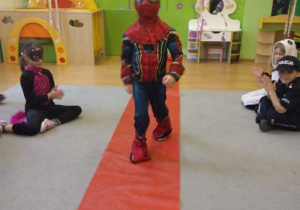 Na podłodze rozłożona jest długa czerwona tkanina. Po niej idzie chłopiec w stroju karnawałowym. Po lewej i po prawej stronie siedzą dzieci. Z tyłu widać zieloną ścianę z kolorowymi naklejkami i meble