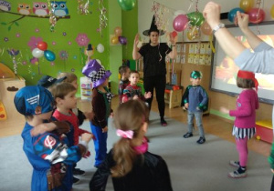 Nauczycielka w czarnym kapeluszu stoi z podniesionymi rękami. Wokół niej stoją dzieci w strojach karnawałowych. W tle widać zieloną ścianę. U góry kolorowe balony i serpentyny.