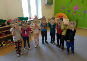 Grupa dzieci stoi w półkolu. Twarze zasłaniają szarymi maskami wyciętymi z papieru. Do każdej maska przyklejone są elementy twarzy: oczy, usta, nos. Za dziećmi widać okno, półkę na zabawki i zieloną ścianę a kolorowymi naklejkami.