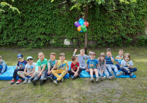Na niebieskim materacu i karimacie siedzi grupa dzieci. W tle na małym drzewku powieszone są kolorowe balony.