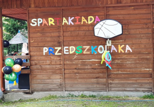 Drewniana ściana domku stojącego w ogrodzie. Na niej zawieszony jest kosz do koszykówki, do którego przyczepione są podłużne poskręcane balony. Na ścianie umieszczono napis "Spartakiada Przedszkolaka".