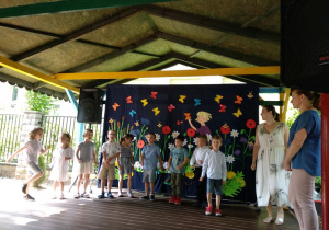 Na scenie pod wiatą ogrodową stoi w półkolu grupa dzieci. Obok dzieci po lewej stronie stoją nauczycielki. Za dziećmi znajduje się dekoracja: wycięte z papieru motyle, kwiaty, postać dziewczynki. Po lewej stronie stoi czarny głośnik.