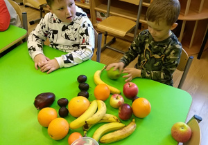 Dwóch chłopców siedzi za stołem. Przed nimi na stole leżą owoce: banany, jabłka, pomarańcze, śliwki, mango oraz pojemnik z truskawkami. Jeden chłopiec trzyma w rękach gruszkę. Za dziećmi widać krzesła i regał z grami w pudełkach.