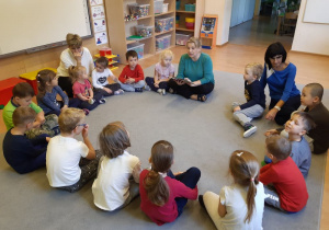 Grupa dzieci siedzi na dywanie w kole. Między nimi siedzą trzy nauczycielki. Jedna z nauczycielek trzyma w rękach książkę. W tle widać półki z zabawkami.