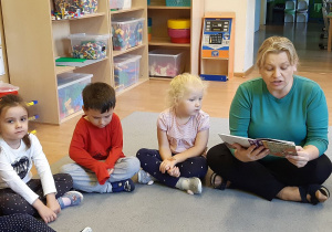 Troje dzieci siedzi obok siebie na dywanie. Z prawej strony siedzi nauczycielka, która czyta książkę. Za nimi widać półki z zabawkami.