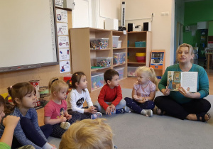 Grupa dzieci siedzi obok siebie na dywanie. Z prawej strony siedzi nauczycielka, która trzyma w rękach odwróconą książkę i pokazuje dzieciom obrazek. W tle widać białą tablicę, półki z zabawkami oraz drugą salę.