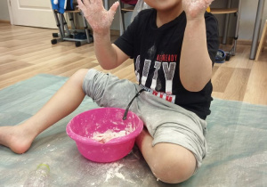 Na podłodze siedzi chłopiec. Chłopiec ma wysunięte dłonie z rozpostartymi palcami, ubrudzonymi mąką. Przed chłopcem stoi różowa miska z białą masą. Za chłopcem widać fragment białych drzwi, tablice i krzesła.