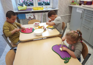Troje dzieci siedzi przy dwóch stolikach. Na jednym stole stoją dwie miski: jedna pusta, druga z owocami oraz talerz z owocami. Dzieci mają przed sobą deski do krojenia, a w rękach noże i kroją owoce. W tle na wprost widać okno, z prawej strony - fragmenty szafek kuchennych