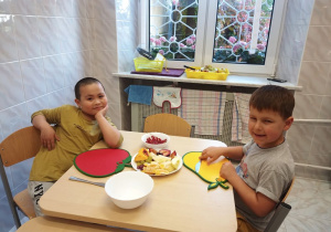 Przy stole siedzą naprzeciwko siebie dwaj chłopcy. Chłopcy patrzą przed siebie i uśmiechają się. Między nimi stoją: pusta miska, miska z czerwonymi owocami i talerz w pokrojonymi na kawałki owocami. W tle widać okno.