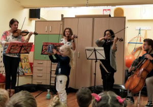 Trzy kobiety stoją obok siebie i trzymają w rękach instrumenty muzyczne: skrzypce i altówkę. Przed każdą kobietą stoi statyw z partyturą. Po lewej stronie siedzi mężczyzna, który trzyma wiolonczelę. Muzycy grają na instrumentach. Przed nimi na środku stoi dziewczynka. Trzyma w ręku metalową puszkę. Z przodu widać głowy dzieci. Z tyłu za muzykami znajdują się szafy.