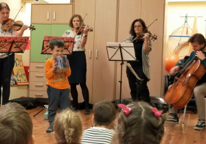 Trzy kobiety stoją obok siebie i trzymają w rękach instrumenty muzyczne: skrzypce i altówkę. Przed każdą kobietą stoi statyw z partyturą. Po lewej stronie siedzi mężczyzna, który trzyma wiolonczelę. Muzycy grają na instrumentach. Przed nimi na środku stoi chłopiec. Trzyma w ręku metalową puszkę. Z przodu widać sylwetki dzieci. Z tyłu za muzykami znajdują się szafy.